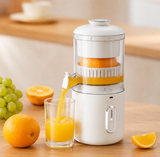 A white countertop electric juicer with a clear upper chamber is shown squeezing an orange and pouring juice into a glass. More oranges, a bunch of green grapes, and a sliced orange are arranged on a wooden table in a bright kitchen setting.